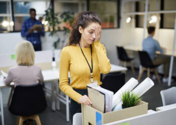 Frustrated young woman in yellow sweater standing at table and touching face with hand while packing stuff in office after dismissal