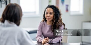 A young woman of mixed race, sits with her female doctor as they discuss her health concerns.  The patient is dressed casually and has a neutral expression on her face.