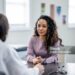 A young woman of mixed race, sits with her female doctor as they discuss her health concerns.  The patient is dressed casually and has a neutral expression on her face.