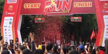 Suasana meriah garis Start dan Finish Alfamart Run 2025 dengan gerbang besar berwarna merah. Terdapat tulisan 'START RUN 2025 FINISH' dan penunjuk waktu untuk kategori 5K dan 10K. Ratusan peserta terlihat mengangkat tangan dan ponsel mereka untuk merekam momen tersebut, di tengah taburan konfeti merah yang berhamburan.