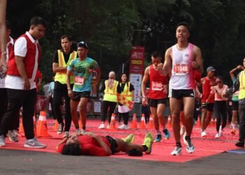 Seorang pelari kelelahan tergeletak di garis finish maraton, sementara pelari lain dan petugas berjaket merah/hijau mengawasi.