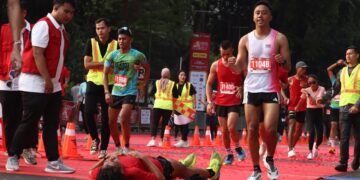 Seorang pelari kelelahan tergeletak di garis finish maraton, sementara pelari lain dan petugas berjaket merah/hijau mengawasi.