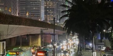 Pemandangan jalanan kota Jakarta di malam hari yang basah karena hujan, dengan gedung pencakar langit, bus, dan lampu kendaraan yang memantul di aspal.