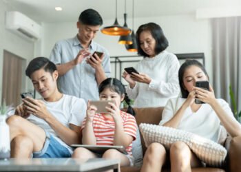 Satu keluarga terdiri dari lima orang duduk dan berdiri di ruang tamu sambil fokus pada ponsel dan tablet masing-masing, menunjukkan bagaimana penggunaan gawai telah menggeser interaksi langsung dalam keluarga.