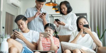 Satu keluarga terdiri dari lima orang duduk dan berdiri di ruang tamu sambil fokus pada ponsel dan tablet masing-masing, menunjukkan bagaimana penggunaan gawai telah menggeser interaksi langsung dalam keluarga.