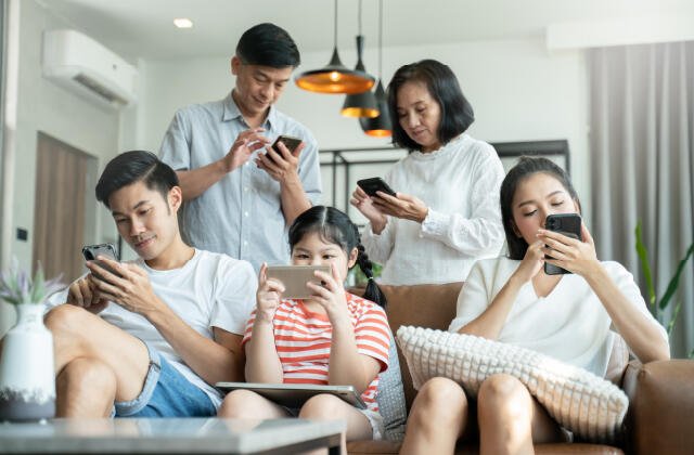 Satu keluarga terdiri dari lima orang duduk dan berdiri di ruang tamu sambil fokus pada ponsel dan tablet masing-masing, menunjukkan bagaimana penggunaan gawai telah menggeser interaksi langsung dalam keluarga.