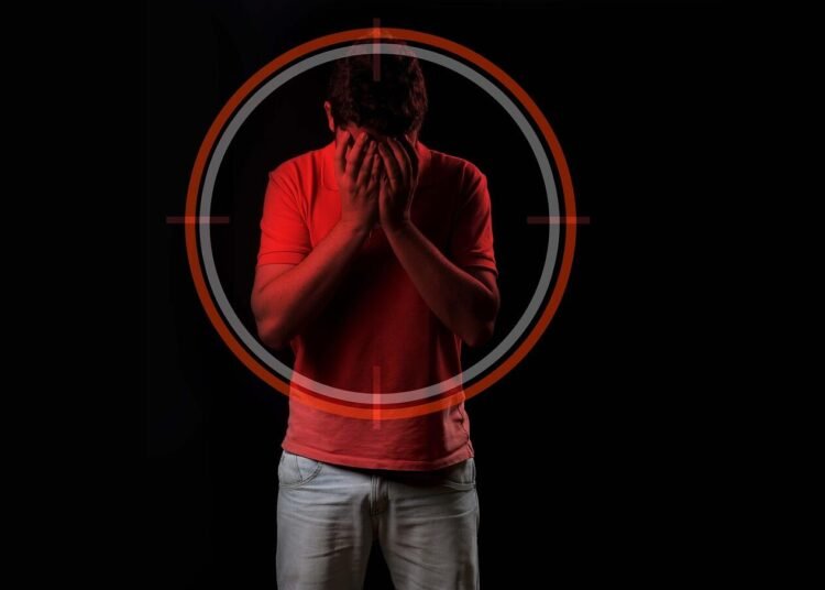 Seorang pria berdiri menunduk sambil menutup wajah dengan kedua tangan, dikelilingi ilustrasi crosshair berwarna merah, menggambarkan tekanan mental atau rasa terperangkap.