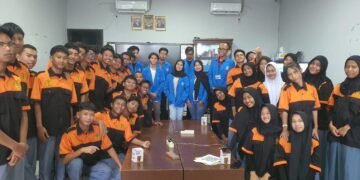 Sebuah foto grup di dalam ruang kelas. Di bagian tengah, delapan mahasiswa PkM UBSI mengenakan jaket almamater biru berbaris dan duduk. Mereka dikelilingi oleh lebih dari dua puluh siswa SMKT Kapin yang mengenakan seragam polo oranye-hitam. Semuanya berpose dan tersenyum ke arah kamera.