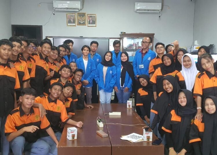 Sebuah foto grup di dalam ruang kelas. Di bagian tengah, delapan mahasiswa PkM UBSI mengenakan jaket almamater biru berbaris dan duduk. Mereka dikelilingi oleh lebih dari dua puluh siswa SMKT Kapin yang mengenakan seragam polo oranye-hitam. Semuanya berpose dan tersenyum ke arah kamera.