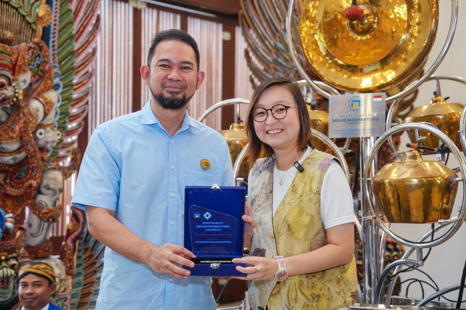 Kementerian Ekraf Luncurkan Robot Gamelan di TMII, Apresiasi Teknologi dan Tradisi 49 Two individuals are smiling and holding a blue plaque in a decorated room with traditional instruments in the background.