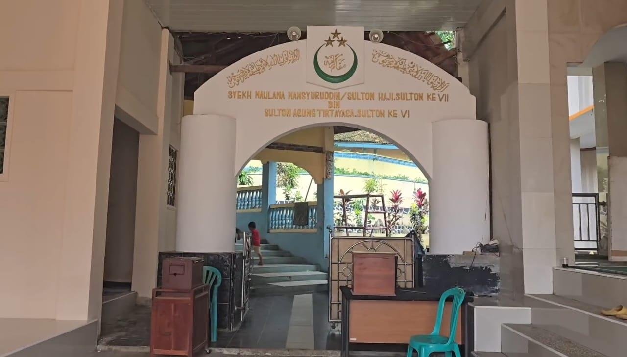 gerbang utama menuju Situs Batu Quran di Pandeglang, Banten adalah sebuah tempat bersejarah yang sarat nilai religius dan budaya. Arsitektur gerbang berbentuk lengkungan dengan dua pilar besar memberi kesan kokoh dan sakral, ditambah ornamen kaligrafi Arab serta lambang khas Banten di bagian atasnya. Suasana ini menandai bahwa pengunjung sedang memasuki area yang dihormati dan dijaga oleh masyarakat setempat. Pada bagian atas gerbang tertulis nama Syekh Maulana Mansyuruddin, seorang tokoh ulama besar sekaligus bangsawan Banten yang memiliki peran penting dalam penyebaran Islam. Penyebutan garis keturunan dari Sultan Agung Tirtayasa menunjukkan bahwa situs ini bukan sekadar tempat ibadah, tetapi juga bagian dari sejarah panjang Kesultanan Banten. Tulisan tersebut memberikan konteks bahwa tempat ini memiliki kaitan kuat dengan perjalanan dakwah dan spiritual ulama terdahulu. Bagian dalam yang tampak dari gerbang menunjukkan area tangga menuju ruang utama situs. Lingkungan yang sederhana namun terawat mencerminkan suasana khas kompleks ziarah. Pengunjung yang datang biasanya menuju ke bagian dalam untuk melihat batu yang dianggap memiliki sejarah unik, berhubungan dengan proses penyebaran ilmu Al-Qur’an oleh Syekh Maulana Mansyur. Pintu gerbang yang menjadi akses menuju salah satu situs religius paling dikenal di Pandeglang. Batu Quran tidak hanya menjadi tempat ziarah, tetapi juga simbol warisan budaya, kesadaran spiritual, dan penghormatan terhadap ulama yang berperan besar dalam membentuk sejarah Islam di Banten.