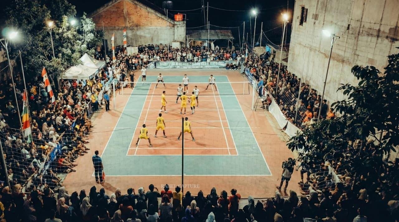 "Tangkapan udara memperlihatkan posisi kedua tim yang tengah bersiap menerima serangan dalam laga Final turnamen Bola Voli IRENA CINANGKA CHAMPIONS II Tahun 2025. Tim berkostum kuning dan tim berkostum putih saling jual beli serangan di tengah kepungan penonton yang memadati seluruh sisi lapangan. Pertandingan yang berlangsung malam hari ini menjadi hiburan utama masyarakat setempat sekaligus ajang pembuktian tim voli terbaik di wilayah Kelurahan Cinangka, Kecamatan Sawangan, Kota Depok."