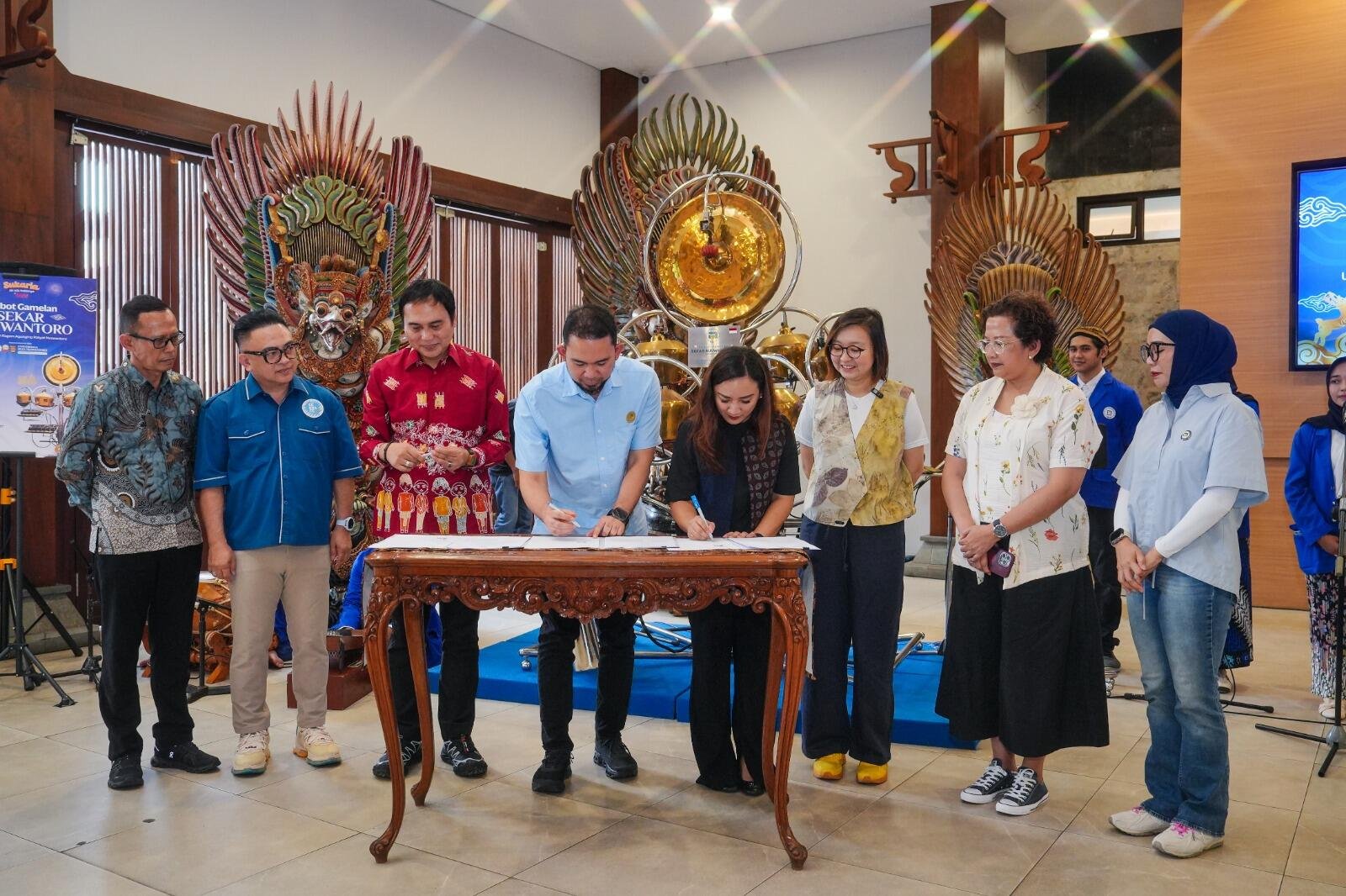 Kementerian Ekraf Luncurkan Robot Gamelan di TMII, Apresiasi Teknologi dan Tradisi 47 A group of people is gathered around a wooden table, signing documents. Traditional decorations and musical instruments are in the background