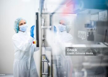 A woman wearing proper safety clothes seen taping on the screen of a big complex machine in pharmaceutical industry while her reflection is visible on the machine.