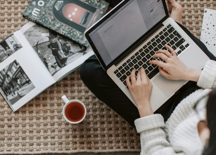 Seorang penulis sedang mengetik "Homework answers" di laptop sambil duduk di lantai, dikelilingi buku, majalah, dan secangkir kopi yang menciptakan suasana produktif dan nyaman.