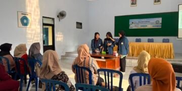 Tiga mahasiswa BBK 8 UNAIR sedang berdiri di depan mendemonstrasikan pembuatan sabun cuci piring kepada ibu-ibu PKK Desa Pilangrejo yang duduk memperhatikan.