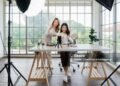Dua wanita Asia membuat konten untuk media sosial. Mereka menggunakan pengaturan studio yang mencakup smartphone yang dipasang di tripod, peralatan pencahayaan, dan berbagai produk riasan yang tersusun di atas meja.
(Sumber: iStock).