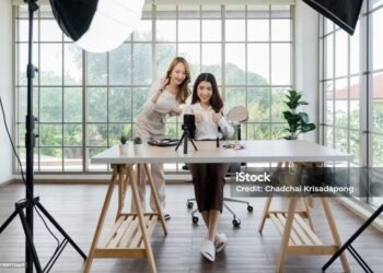 Dua wanita Asia membuat konten untuk media sosial. Mereka menggunakan pengaturan studio yang mencakup smartphone yang dipasang di tripod, peralatan pencahayaan, dan berbagai produk riasan yang tersusun di atas meja.
(Sumber: iStock).