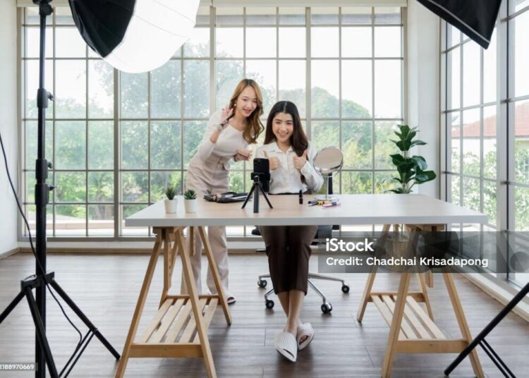 Endorse Bisa Dibayar, Kepercayaan Tidak: Ujian Keaslian dalam Influencer Marketing 46 Dua wanita Asia membuat konten untuk media sosial. Mereka menggunakan pengaturan studio yang mencakup smartphone yang dipasang di tripod, peralatan pencahayaan, dan berbagai produk riasan yang tersusun di atas meja.
(Sumber: iStock).
