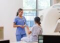 Keterangan foto: Ilustrasi radiografer perempuan yang bekerja menggunakan peralatan radiologi dengan menerapkan prosedur keselamatan radiasi untuk melindungi diri dari paparan radiasi saat bertugas. {Sumber: iStock).